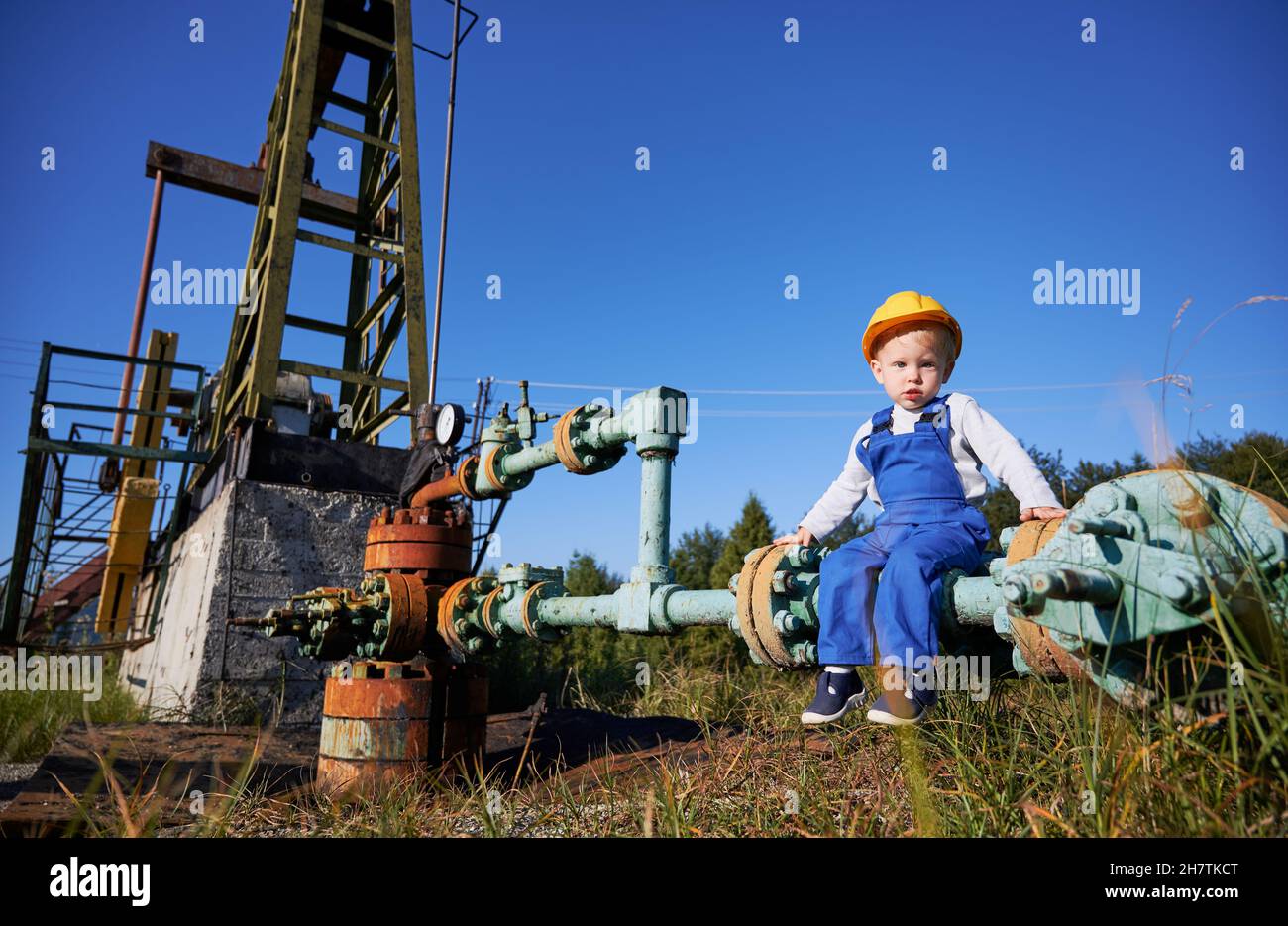 Cute child in construction helmet sitting on pipe of petroleum pump jack under beautiful blue sky. Adorable little boy in work overalls resting on pipe near oil pump rocker-machine in oil field. Stock Photo