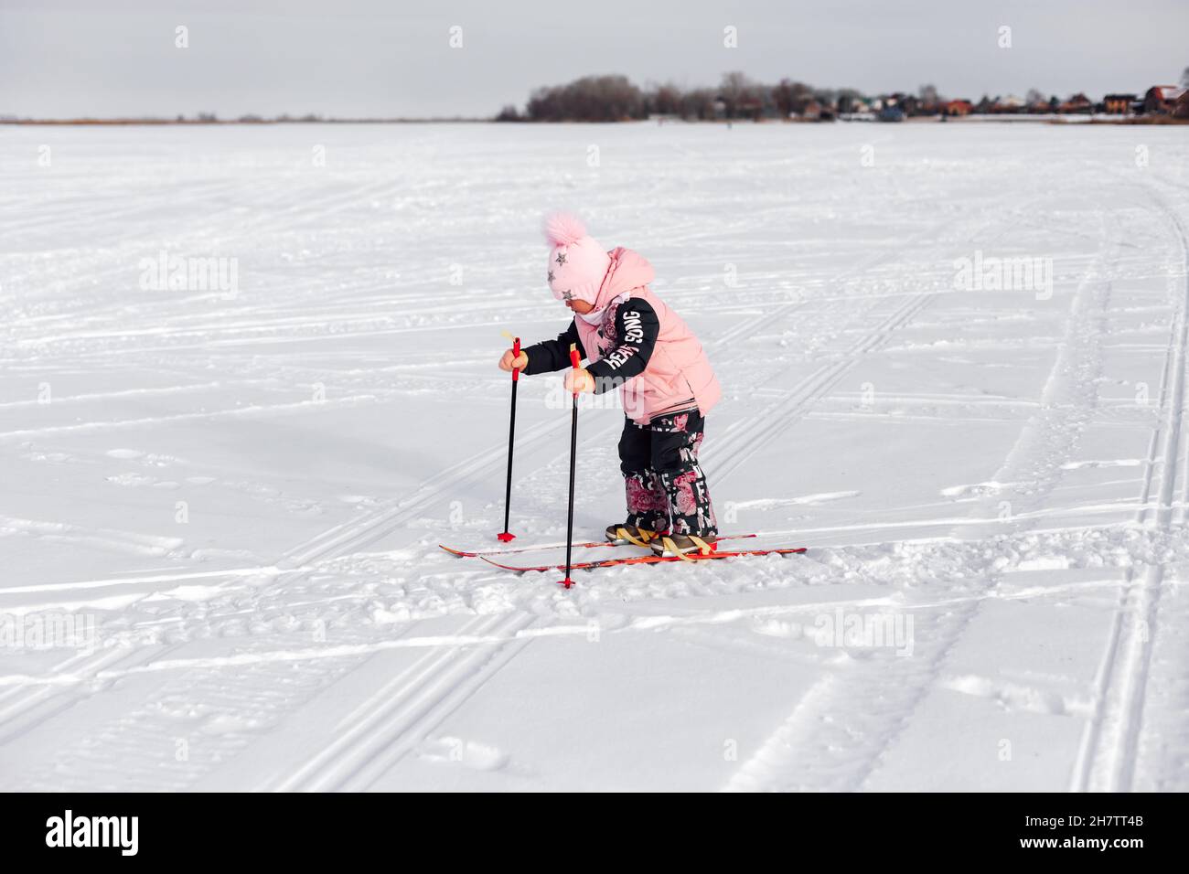 Girl is learning to ski. Little child in pink suit is skiing, she is standing on snowy road on background of winter forest, side view Stock Photo