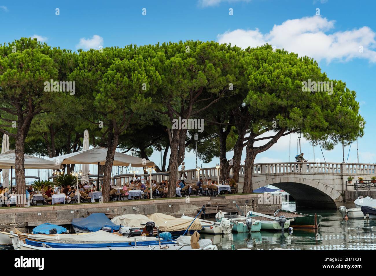 Desenzano, Italy-October 2021; Close up view of people on a terrace under large pine trees in the inner harbor Porto Vecchio with numerous small fishi Stock Photo