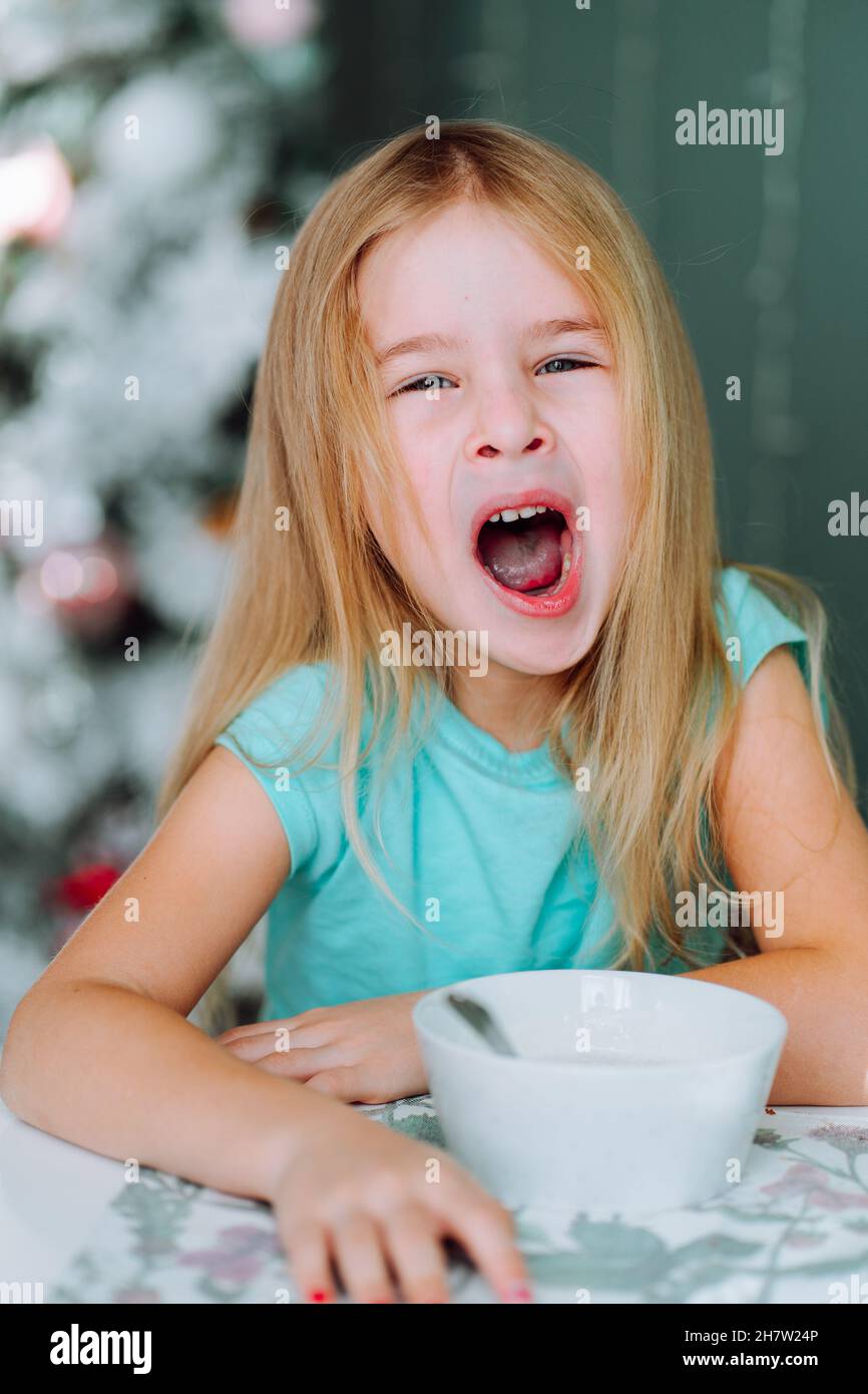 Portrait of yawning little blonde girl having breakfast at morning at winter time with christmas tree on background. Lack of sleeping, high school loa Stock Photo