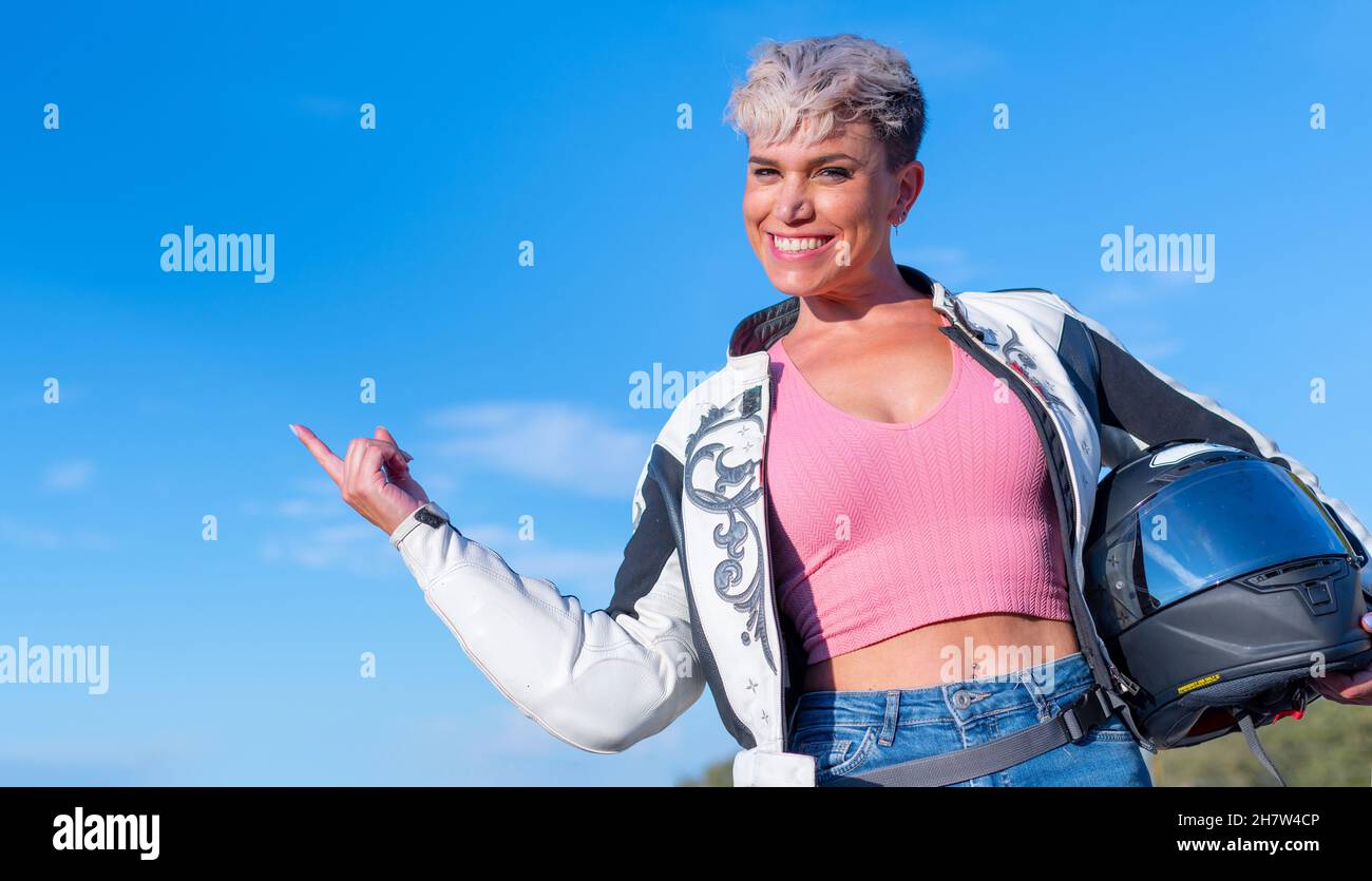 Young pretty blonde woman with short hair dressed as a biker in a fur jacket and safety helmet points up with her finger on a blue sky background Stock Photo