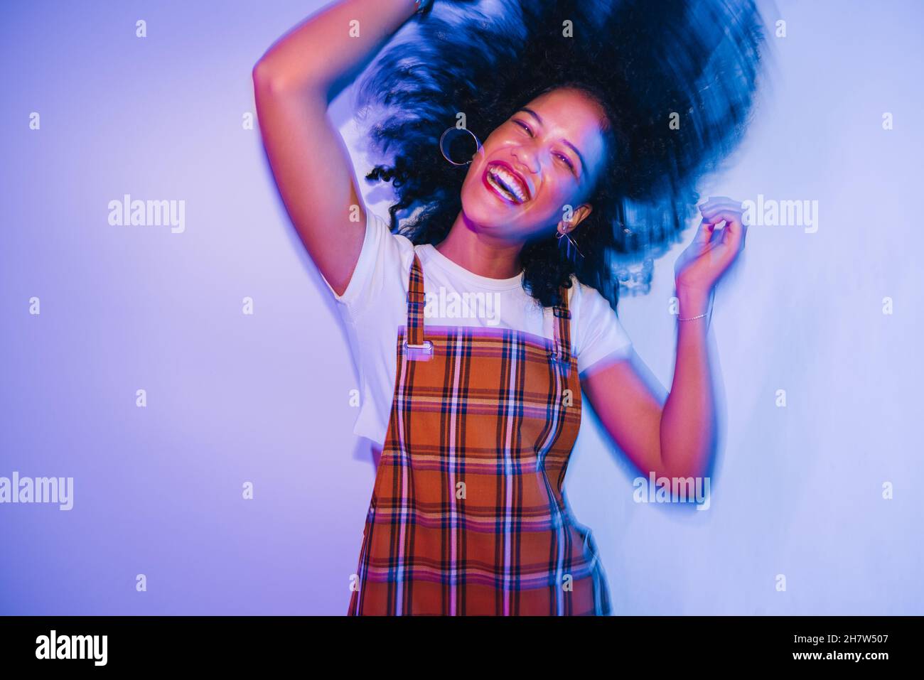 Energetic young woman dancing and whipping her hair happily. Cheerful young woman laughing joyfully while standing alone in bright neon light. Carefre Stock Photo