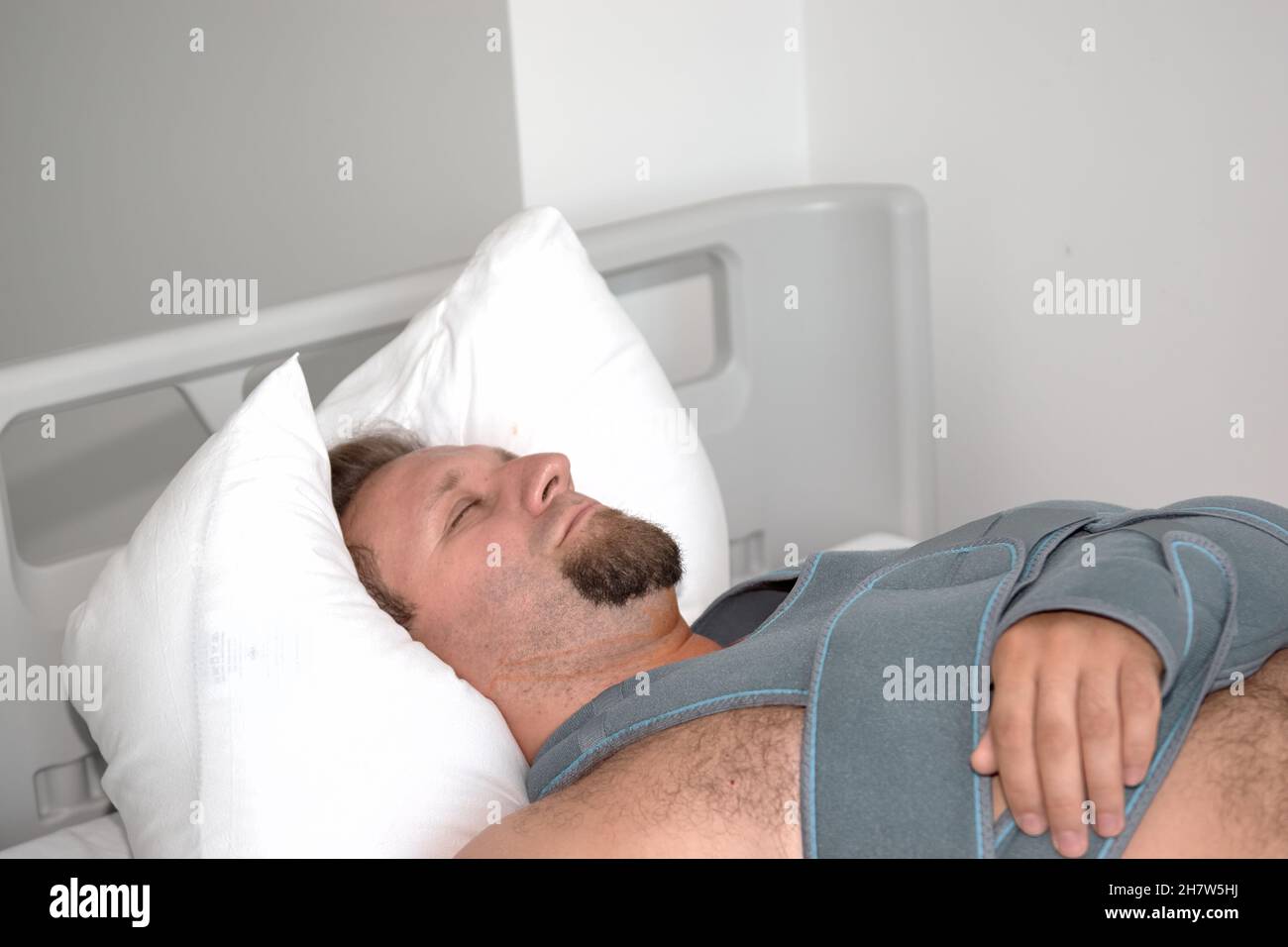 A patient young man with dropper catheter lying on bed in hospital ward. Traumatology Surgery medical procedure in the emergency. Elastic bandage shoulder fracture Stock Photo