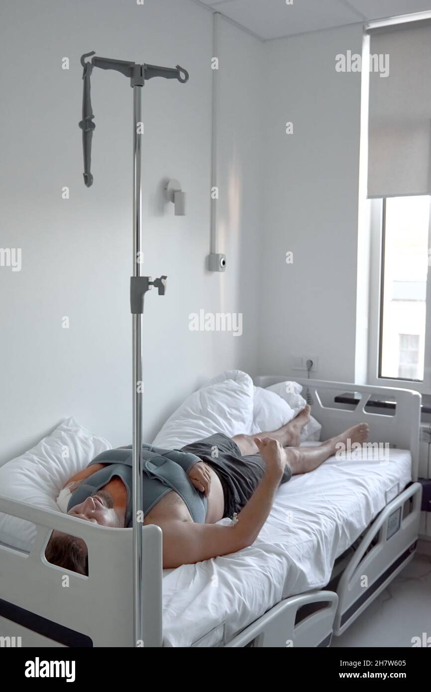 A patient young man with dropper catheter lying on bed in hospital ward. Traumatology Surgery medical procedure in the emergency. Elastic bandage shoulder fracture Stock Photo