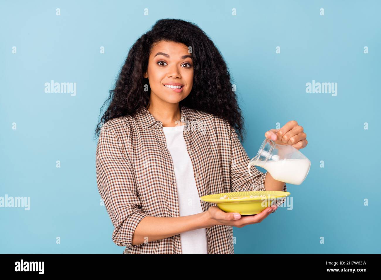 Photo of sweet guilty dark skin lady wear plaid shirt cooking milk cornflakes biting lip isolated blue color background Stock Photo