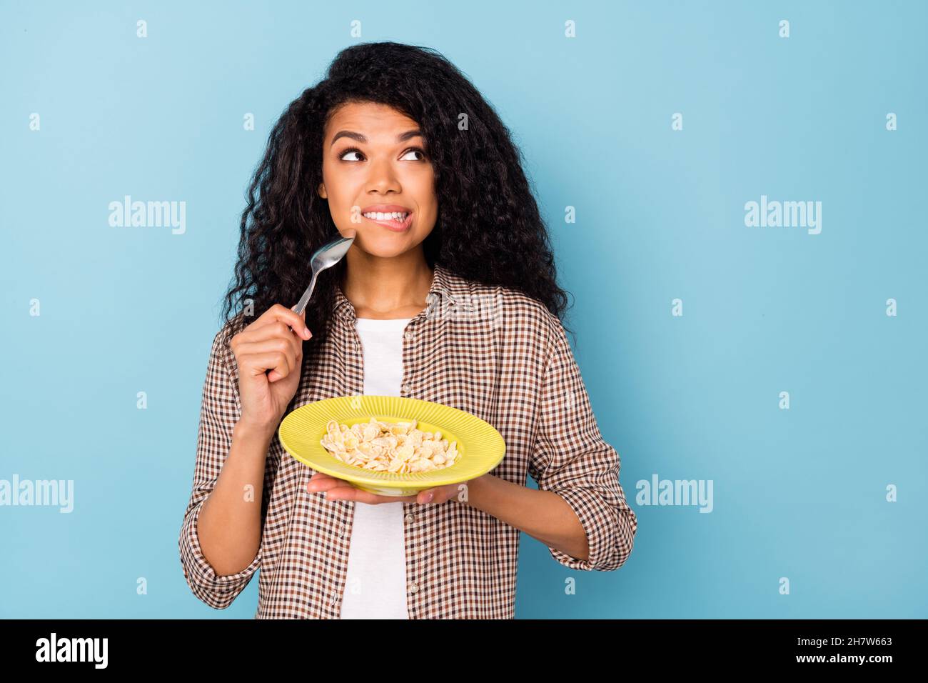 Photo of hungry doubtful dark skin woman dressed shirt eating cornflakes looking empty space isolated blue color background Stock Photo