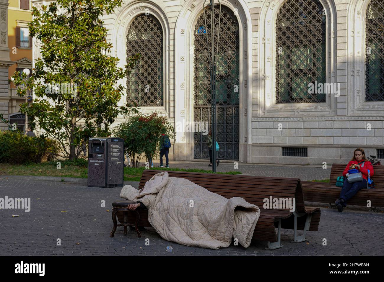 A homeless man in his sleeping bag lies asleep on a bench in front of a