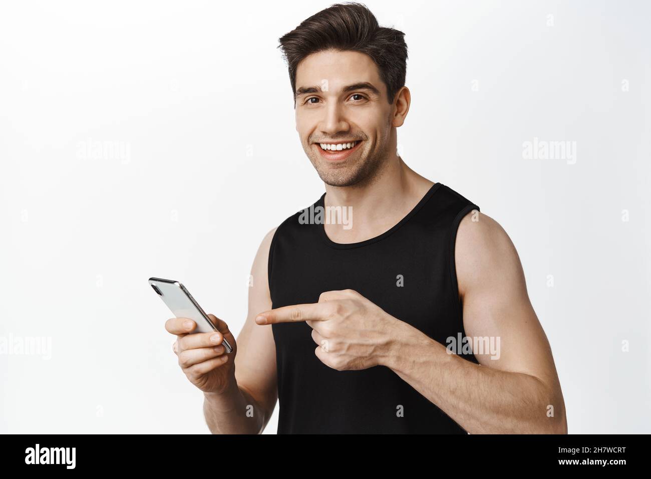Handsome man pointing at his smartphone, doing workout, sport application while attending gym, standing over white background Stock Photo