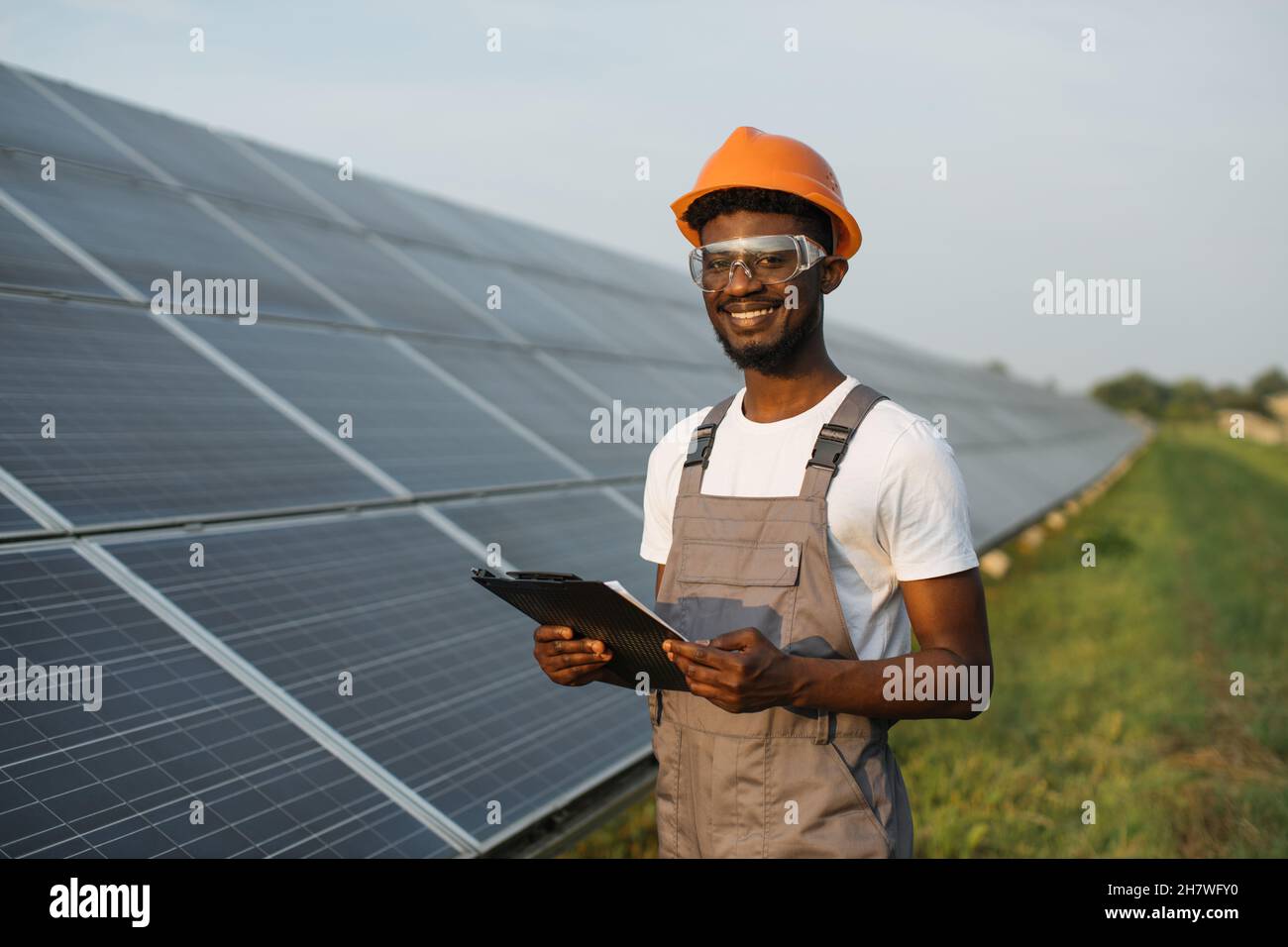 Portrait of smiling african american man in safety glasses and orange helmet holding clipboard while standing on field with solar panels. Concept of service work and alternative energy. Stock Photo