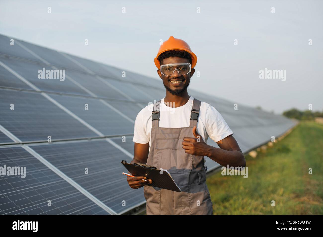Portrait of african american man in safety glasses and orange helmet holding clipboard while standing on field with solar panels showing thumb up. Concept of eco energy and service work Stock Photo
