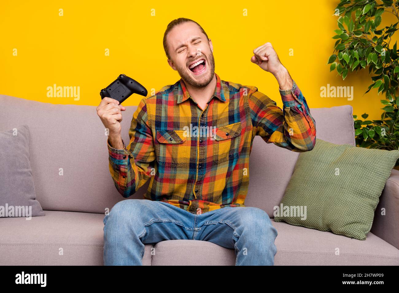 Photo of cheerful astonished guy sit divan play video game rejoice goal wear plaid shirt isolated yellow color background Stock Photo