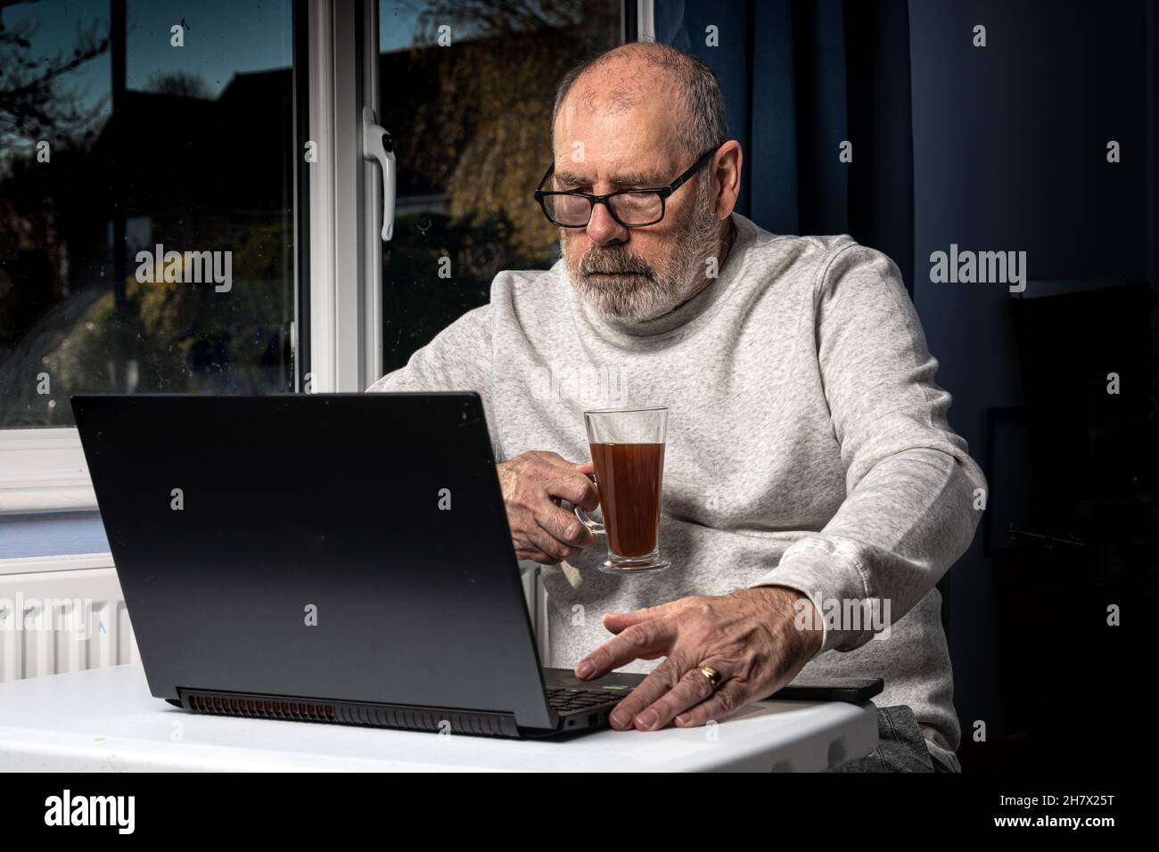 Grey bearded man drinking coffe whilst working on a laptop from his home office. Stock Photo