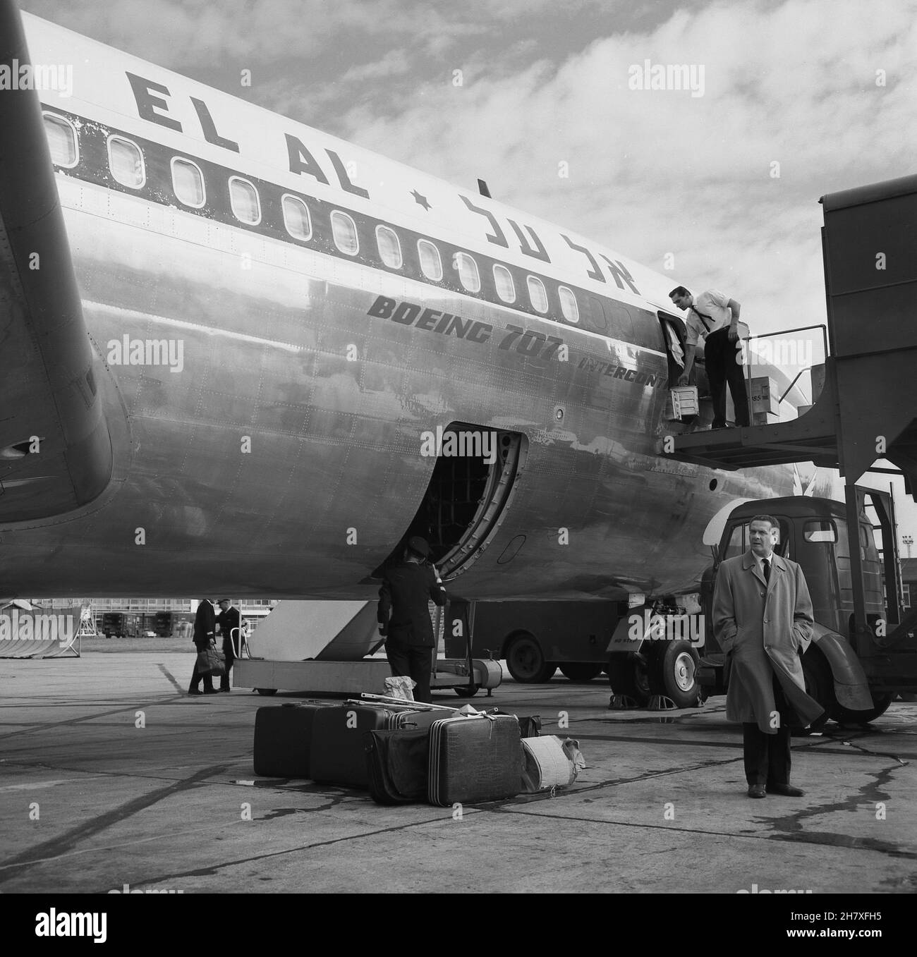 1960s, historical, an EL AL Boeing 707 jet aircraft parked at London Airport, with luggage on the ground and a man in a raincoat standing by it, London, England, UK. A commissary agent of the airline is up on the opened door of a container, loading goods or galleys onto the rear of the aircraft. Stock Photo