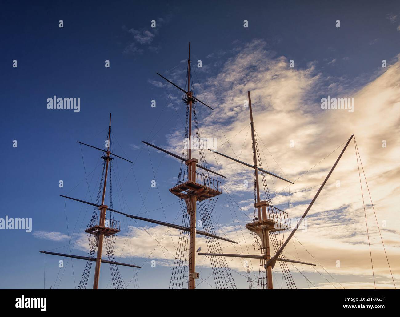 The masts of a tall ship are silhouetted against a sky with light cloud ...