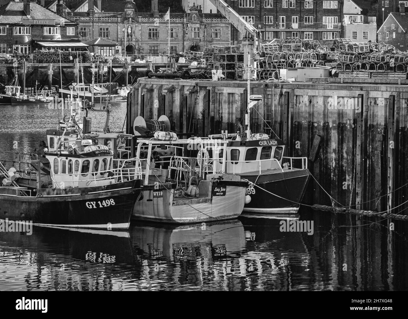 Small fishing boats are moored alongside a quay and buildings are in the background. Lobster pots are stacked on the wharf. Stock Photo