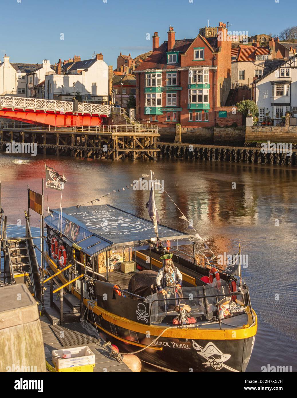 A tour boat on a pirate theme is moored to a quay.  A bridge is in the background and buildings are on the far side of the harbour. A clear sky is abo Stock Photo