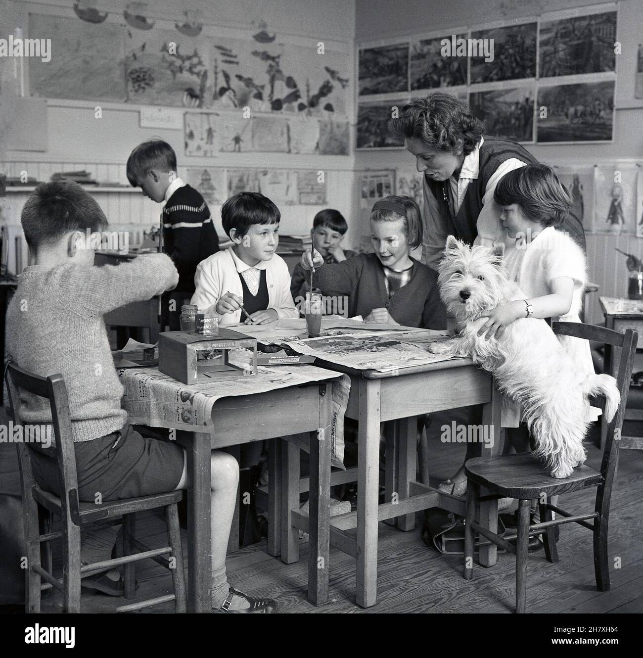 1965, historical, in a primary school classroom, several young children sititing at traditional wooden desks covered with old newspaper doing painting, supervised by a lady teacher. Beside them, a small girl holding the school dog, a West Highland terrier, commonly known as a Scottie, who is standing with its back legs on a chair and its front ones on a desk, Fife, Scotland, UK. Stock Photo