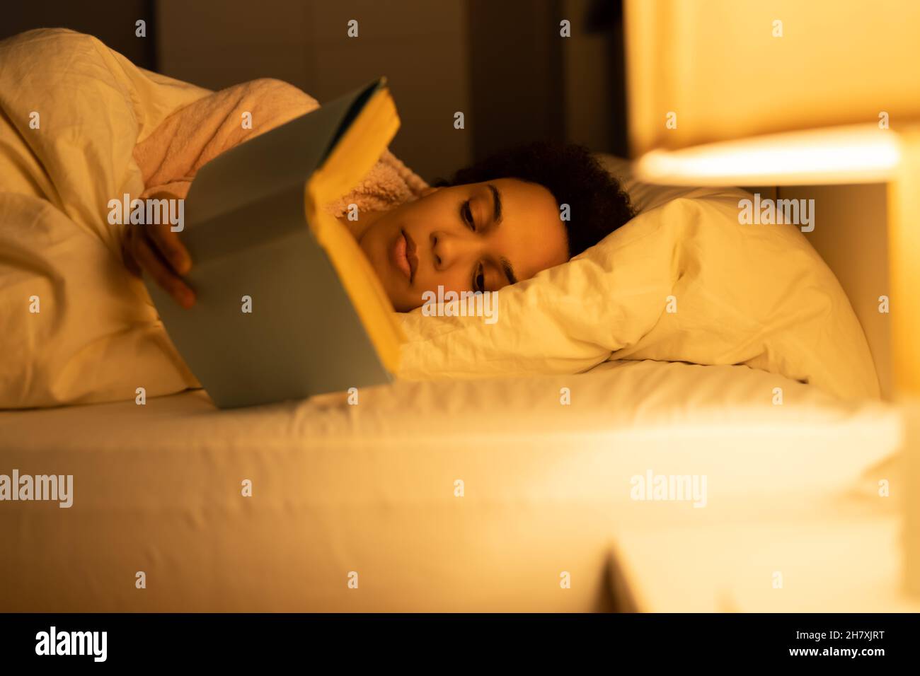 African American Woman Reading Book In Bed At Night Stock Photo