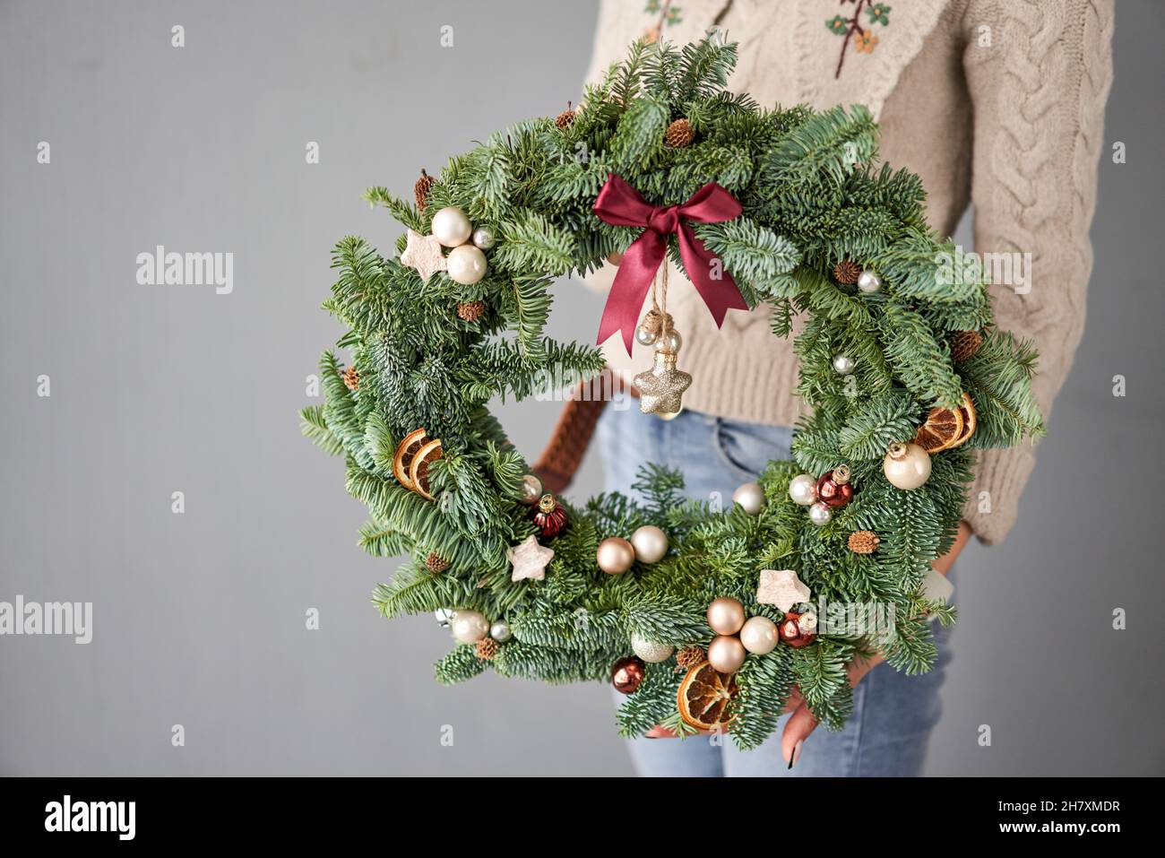 Christmas wreath of fresh spruce in woman hands. Xmas circlet with ornaments, toys and balls. Christmas mood. Gray wall on background Stock Photo