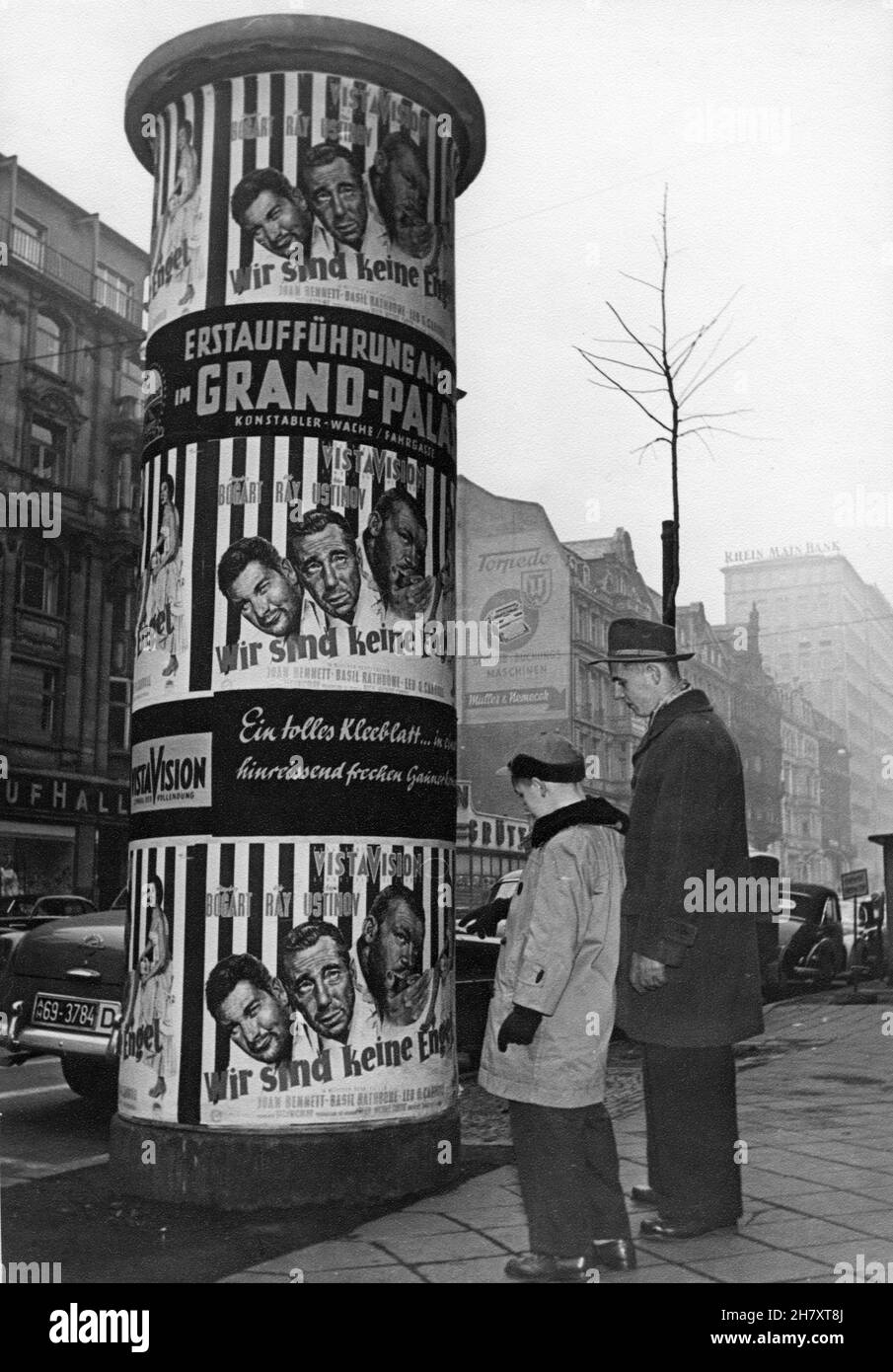 Father and Son on Kaiserstrasse in Frankfurt West Germany in November 1955 looking at Poster Column advertising HUMPHREY BOGART ALDO RAY and PETER USTINOV in WE'RE NO ANGELS 1955 director MICHAEL CURTIZ play Albert Husson costume design Mary Grant Paramount Pictures Stock Photo