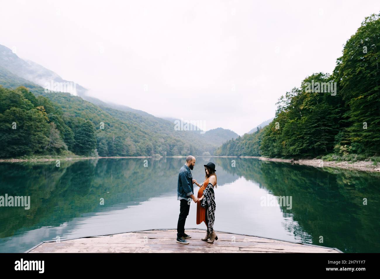 Pregnant woman and man stand holding hands on the pier by the lake ...