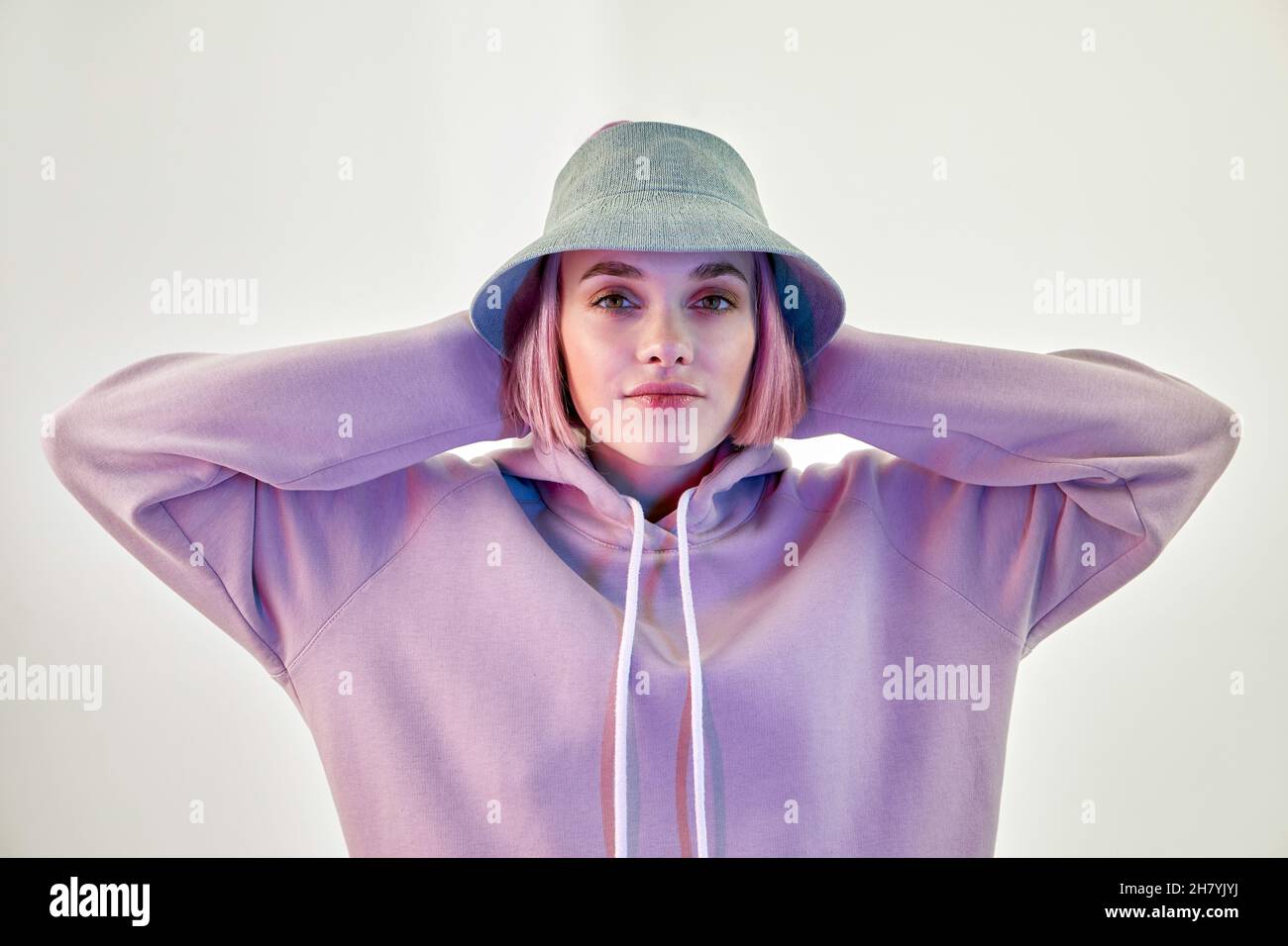 Self assured young female millennial with pink hair in trendy panama hat and hoodie touching head and looking at camera against white background Stock Photo