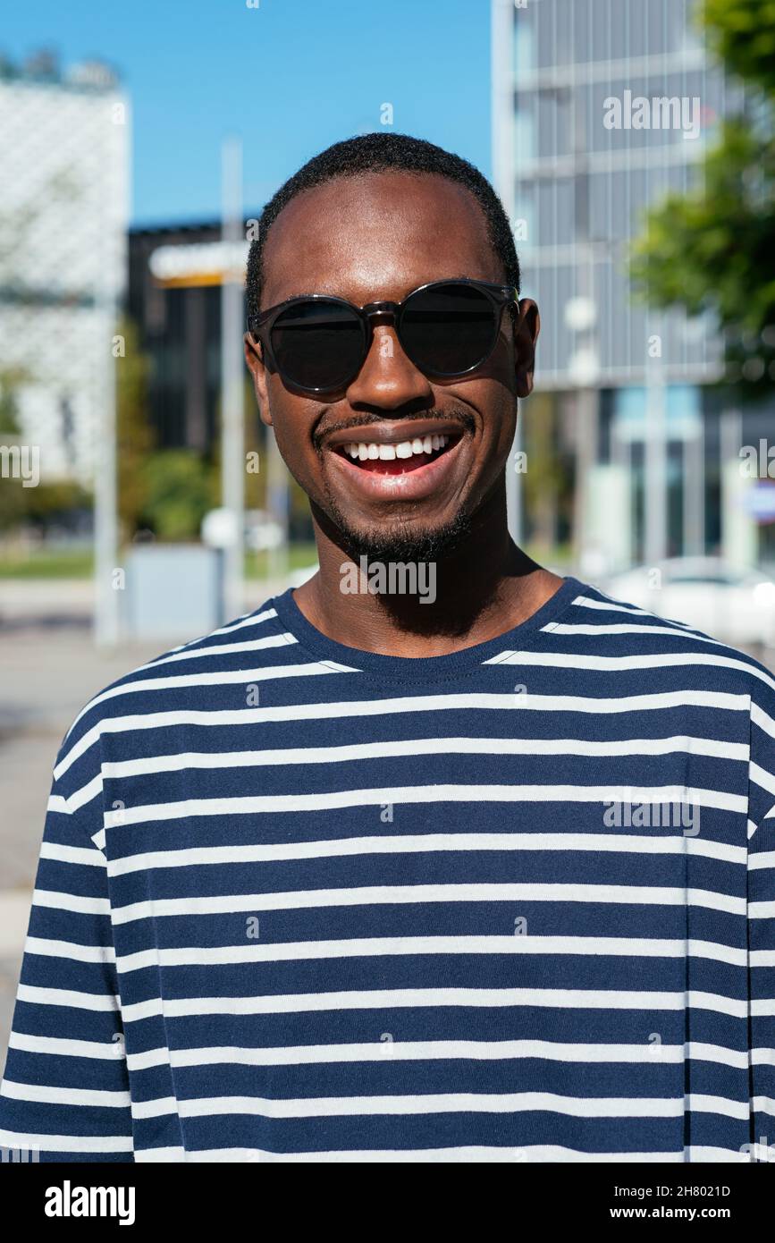Optimistic African American male in stylish sunglasses and striped shirt standing on street with modern building in city on sunny day Stock Photo