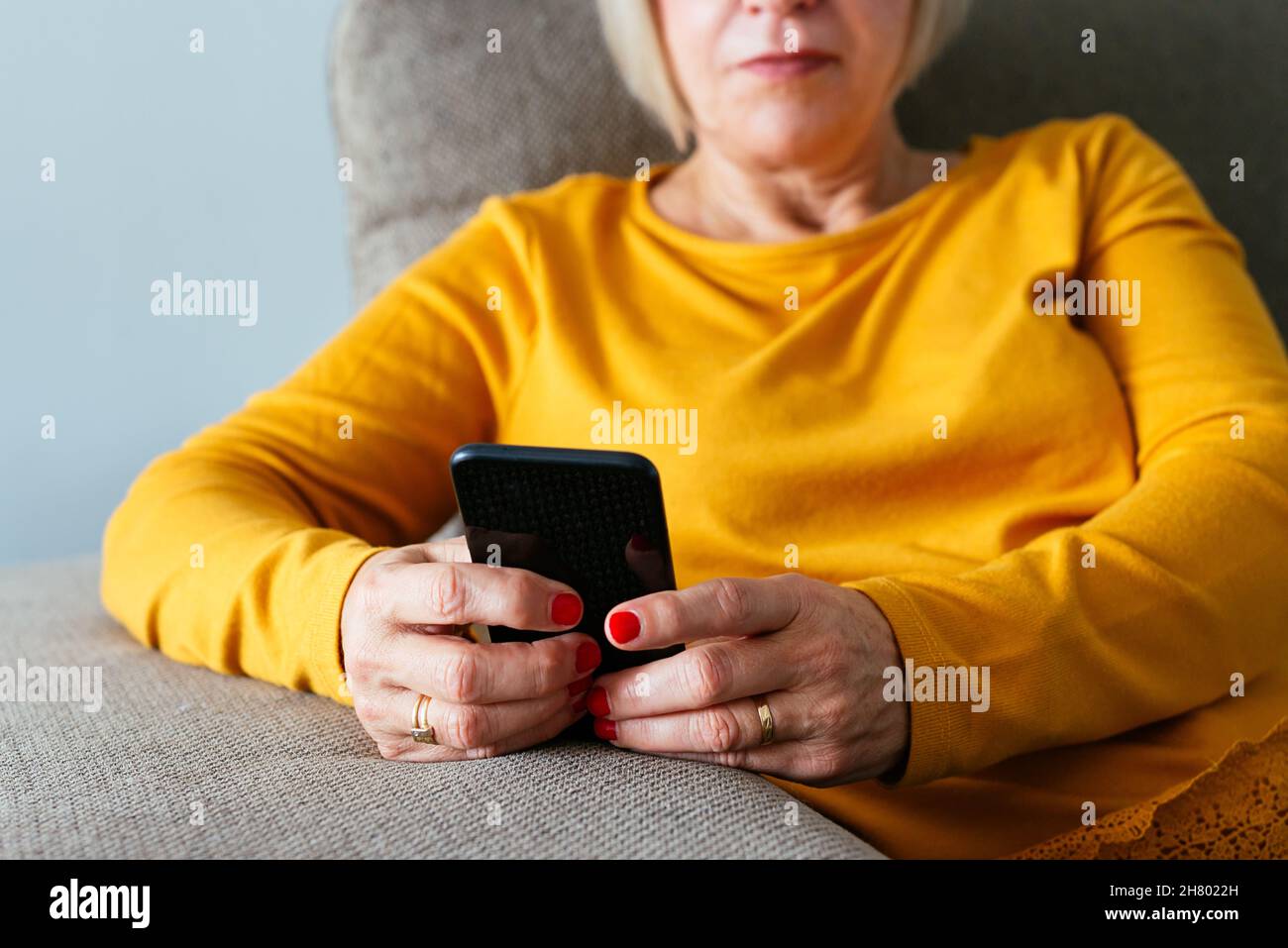Anonymous middle aged female sitting on sofa and browsing mobile phone at home Stock Photo