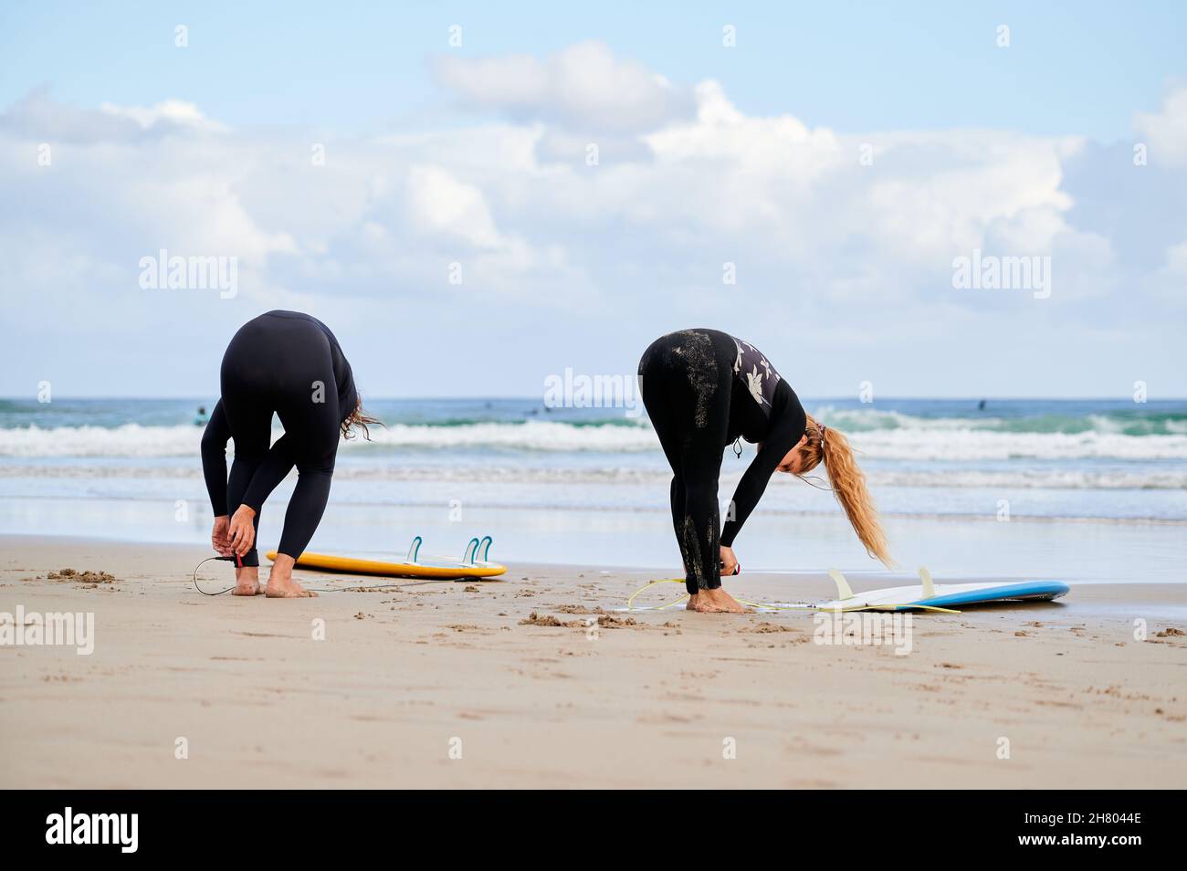 Full body back view of female surfers in wetsuits fastening ankle
