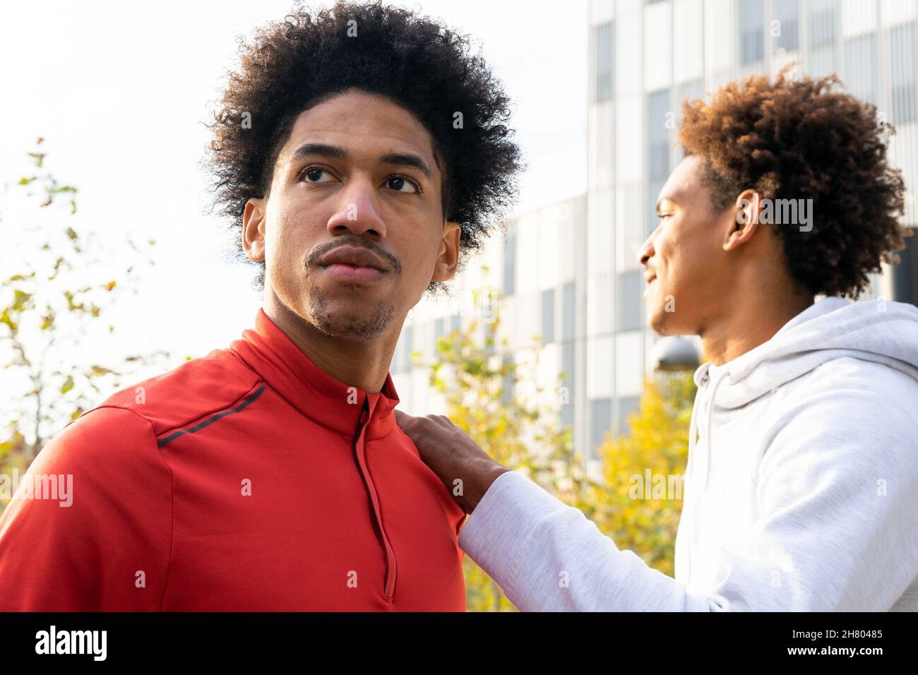 African American man with curly hair putting hand on shoulder of friend on street near building and trees Stock Photo