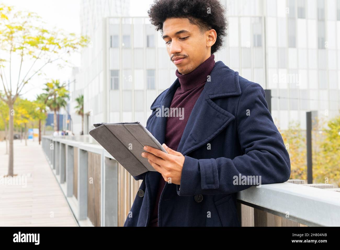 Positive African American male with curly hair dressed in coat browsing on tablet while smiling on street Stock Photo