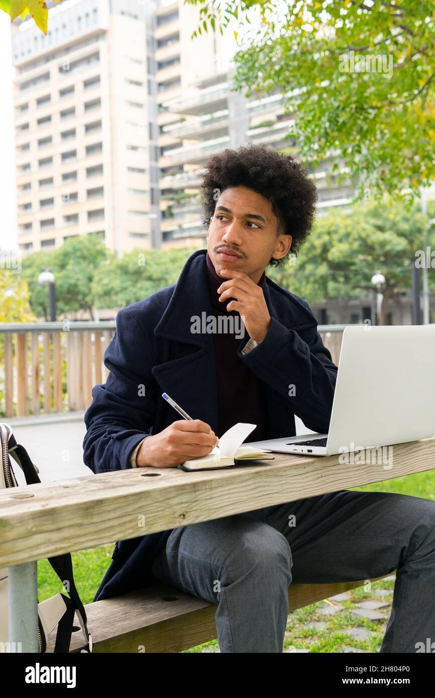 Pensive African American male student in outerwear sitting on bench and taking notes in notepad while doing homework on netbook in park Stock Photo