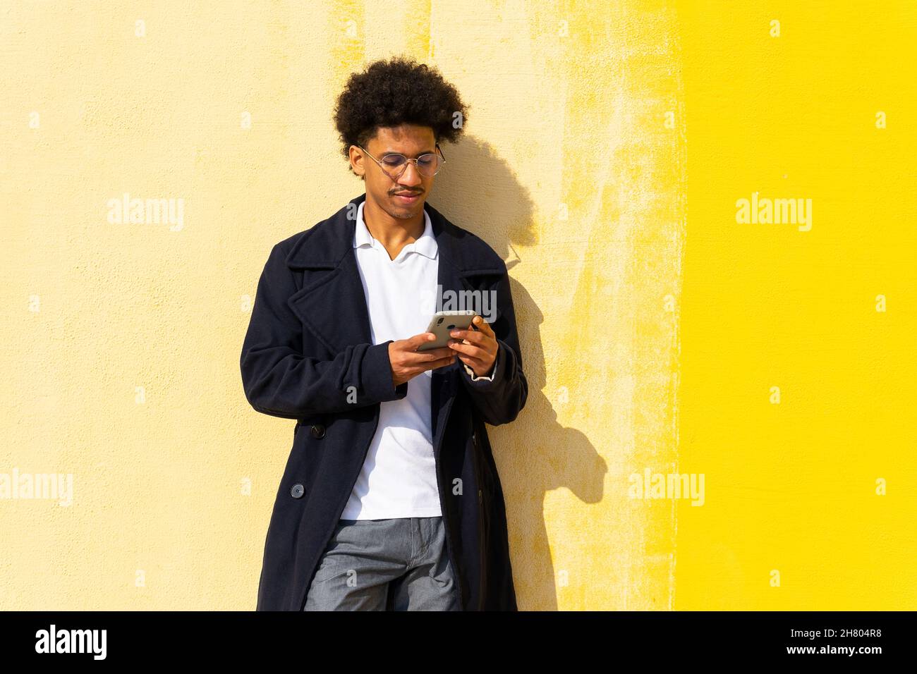 Happy African American male browsing on cellphone while leaning against yellow wall Stock Photo