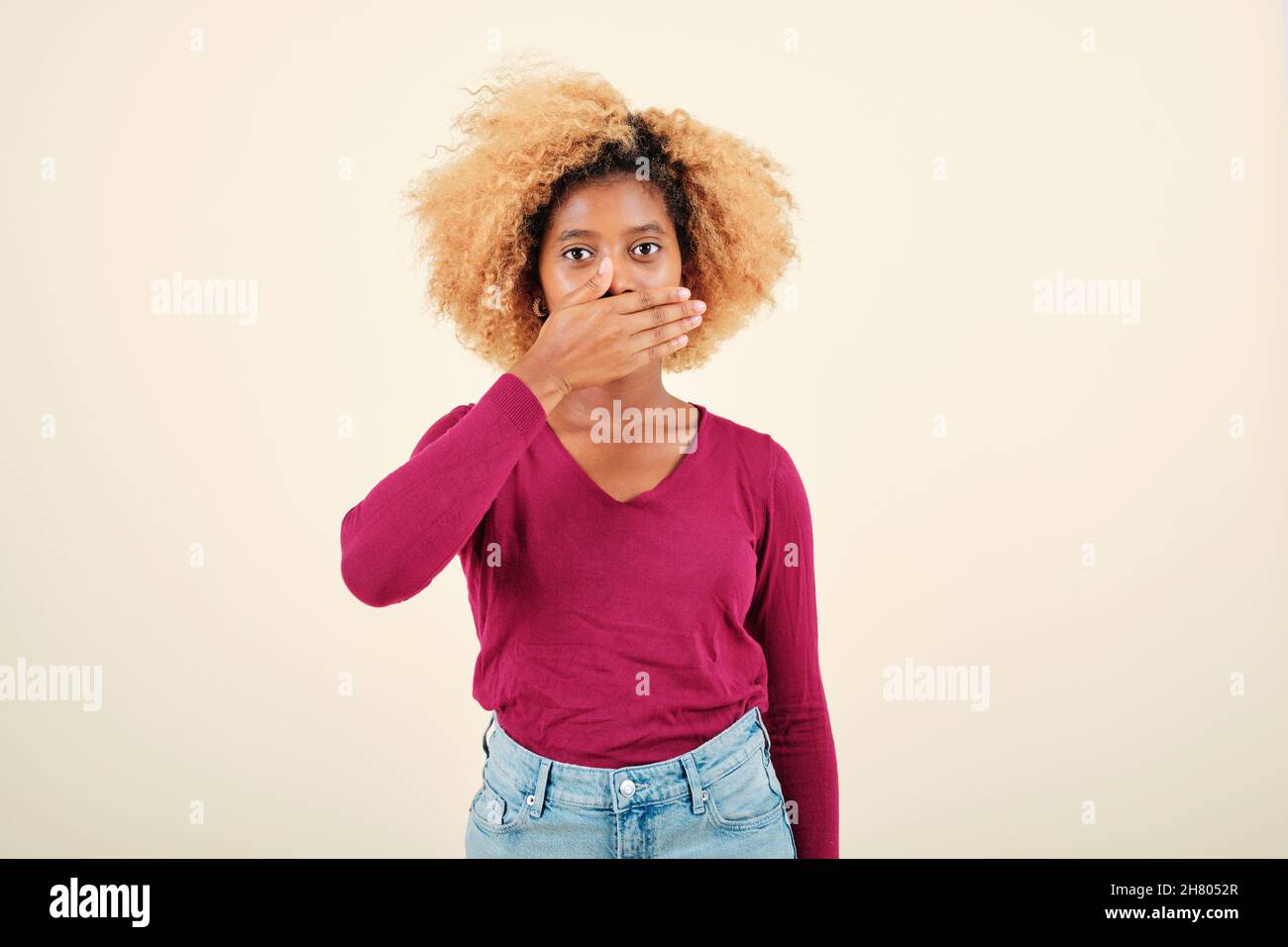 Young woman with afro hairstyle looking shocked while covering mouth with hands. Stock Photo