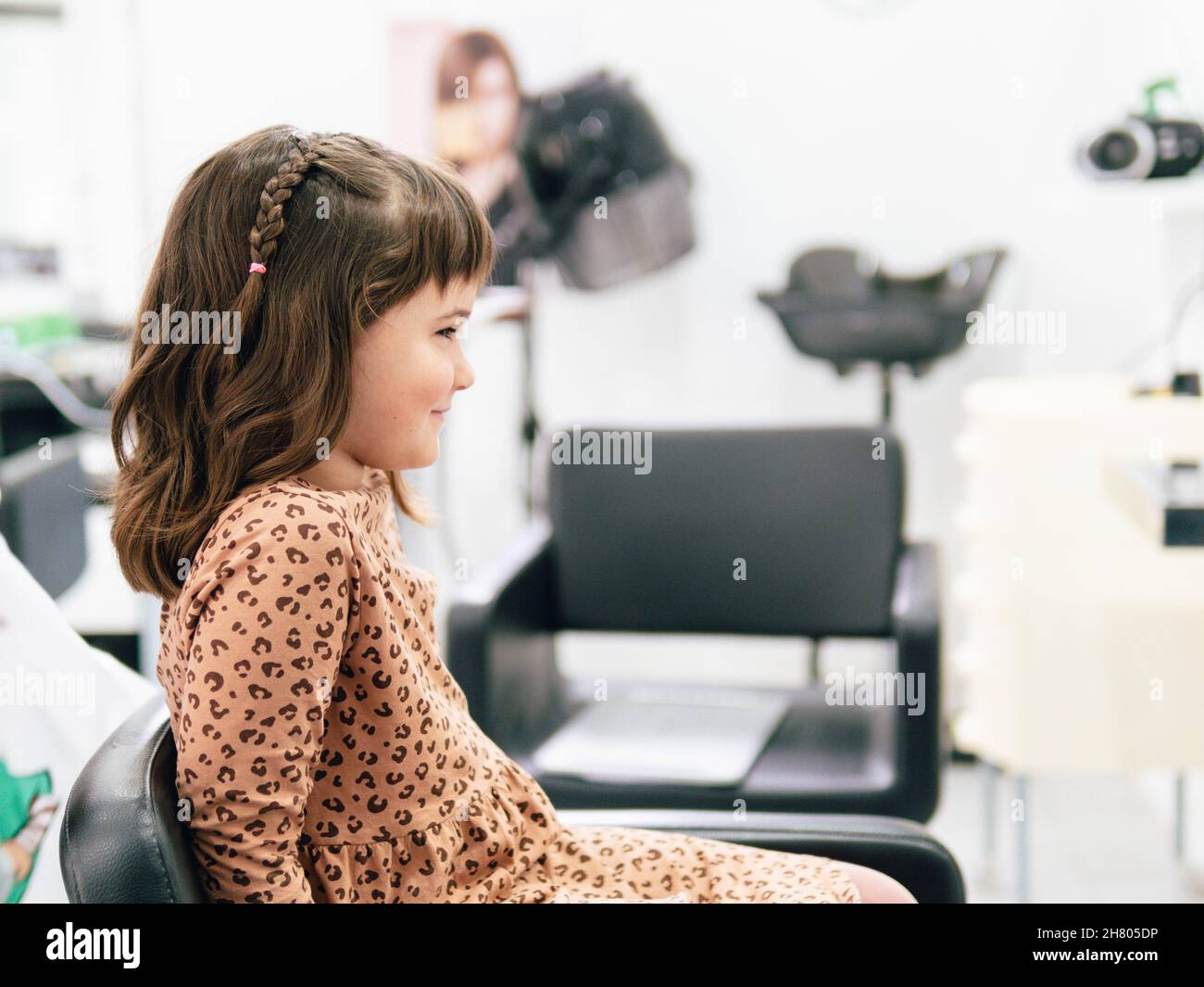 Side view of cute little girl with pigtail and wavy hair sitting in armchair in light modern hairdresser shop Stock Photo