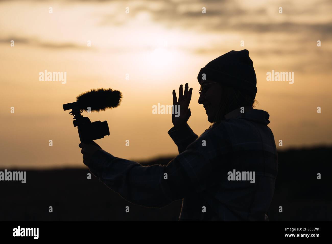 Side view silhouette of unrecognizable young female blogger in hat smiling and showing waving hand while shooting video on digital camera against suns Stock Photo