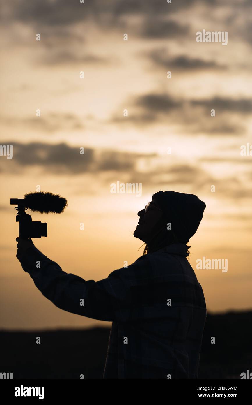 Side view silhouette of unrecognizable young female blogger in hat shooting video on digital camera against sunset sky in countryside Stock Photo