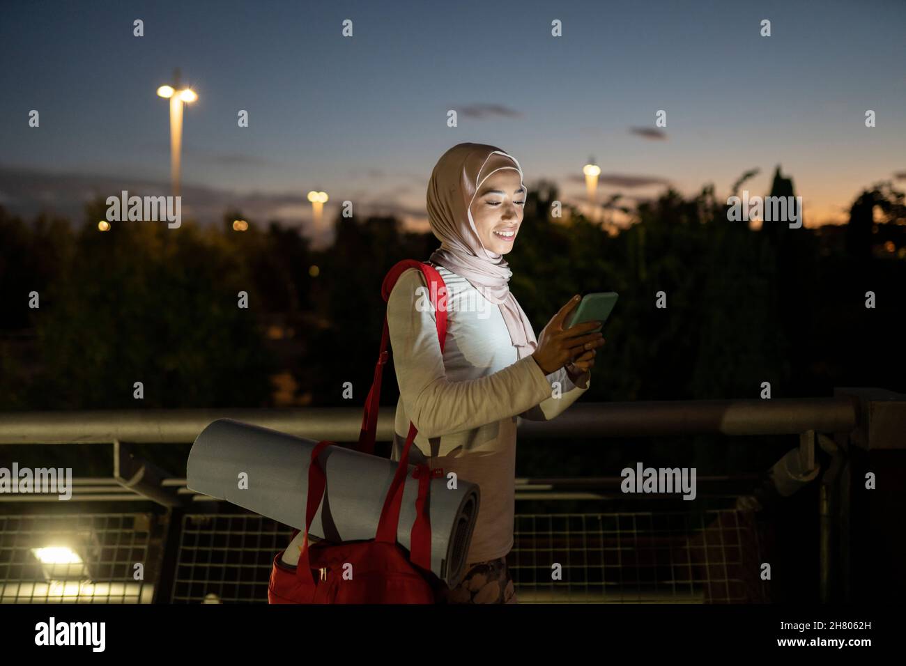 Positive Islamic female in traditional headscarf with sports mat on bag browsing cellphone after training at dark sunset Stock Photo