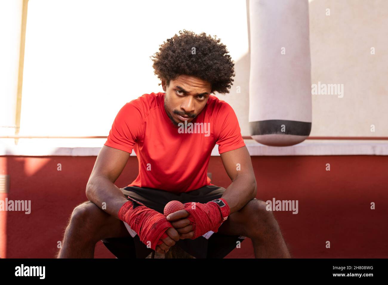 Confident African American sportive man in boxing bandages and activewear looking at camera while sitting near punching bag on street Stock Photo