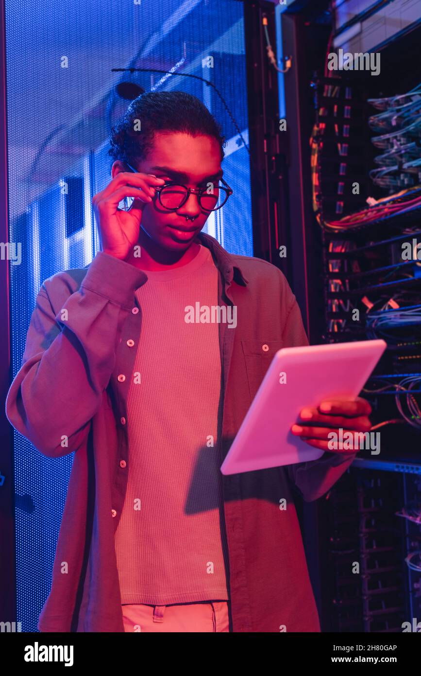 young african american programmer adjusting eyeglasses while looking at digital tablet in data center Stock Photo