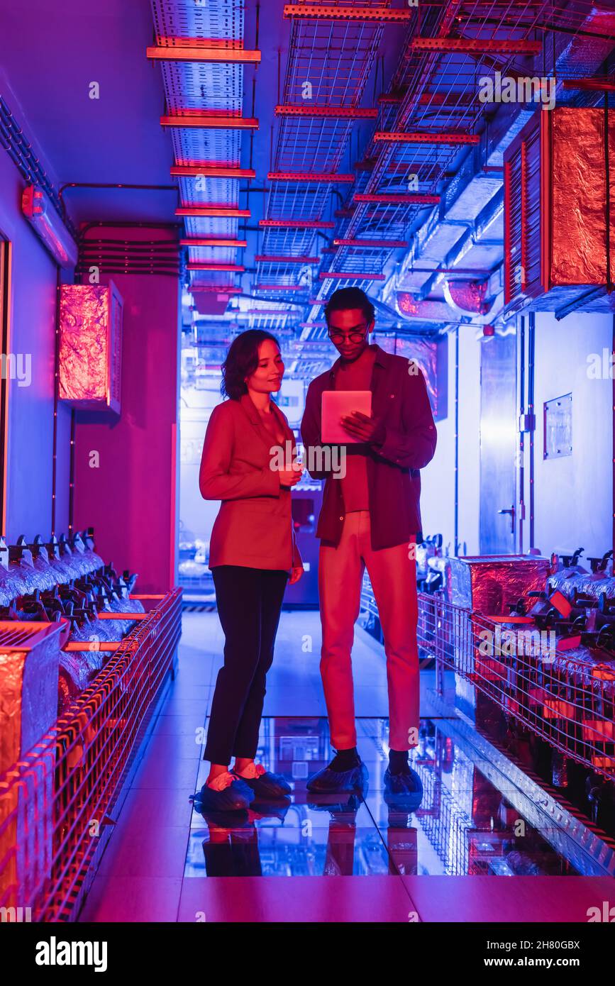 interracial technicians looking at digital camera while standing in technical equipment room in neon light Stock Photo
