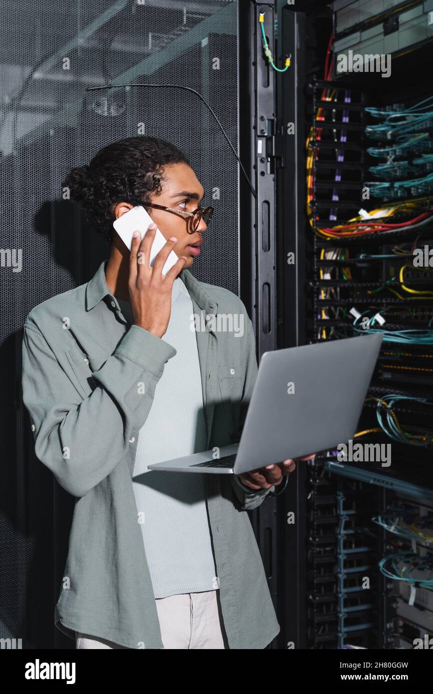 african american technician with laptop talking on smartphone while checking server in data server Stock Photo