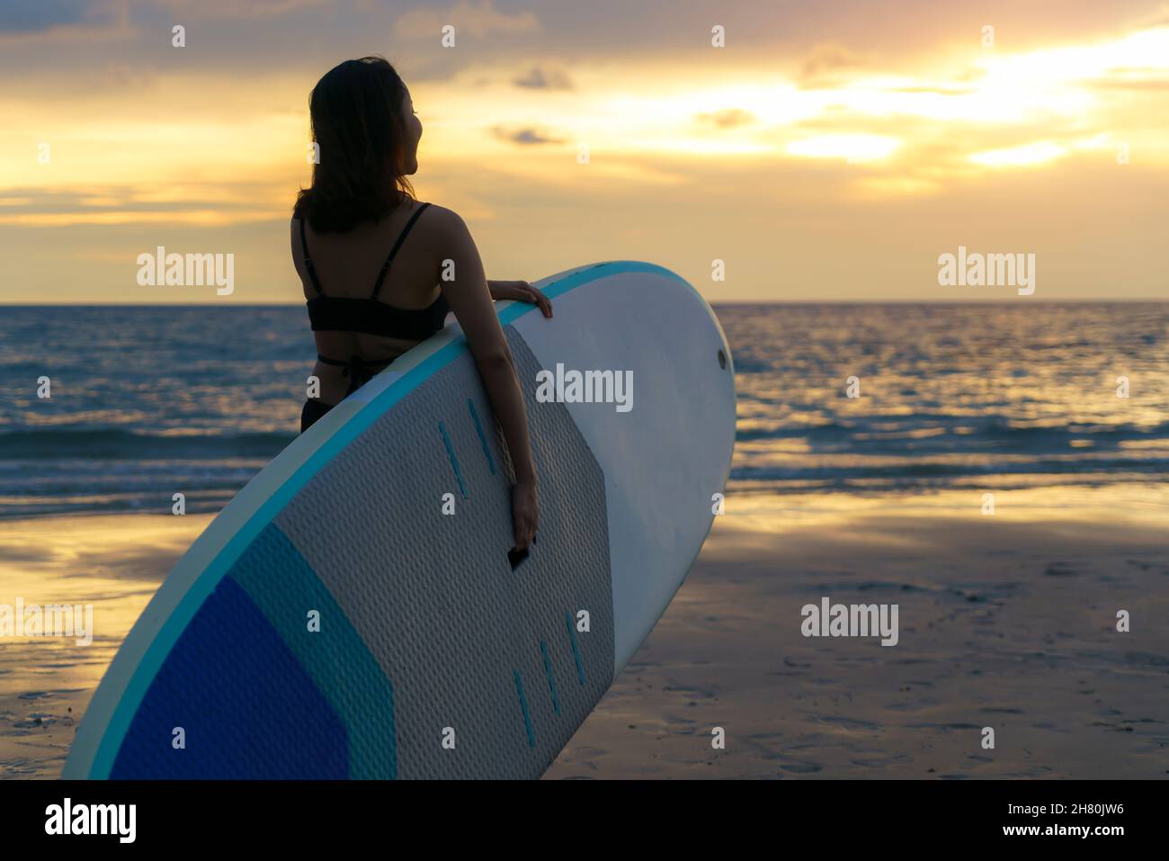 Asian woman holding a sup board and paddle and walking on the beach. In the background, the ocean and the sunset. Back view. Summer surfing. Stock Photo