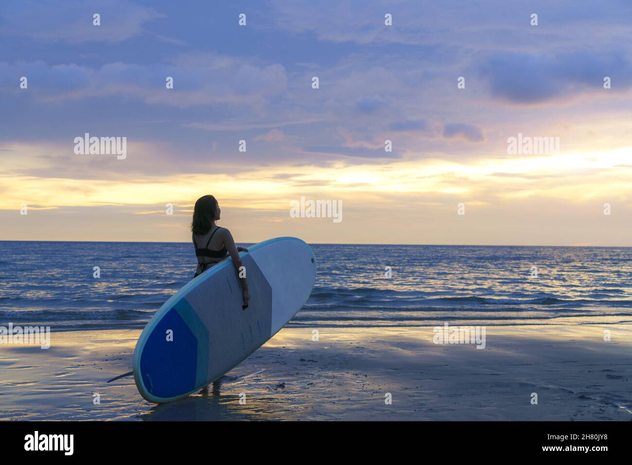 Asian woman holding a sup board and paddle and walking on the beach. In the background, the ocean and the sunset. Back view. Summer surfing. Stock Photo