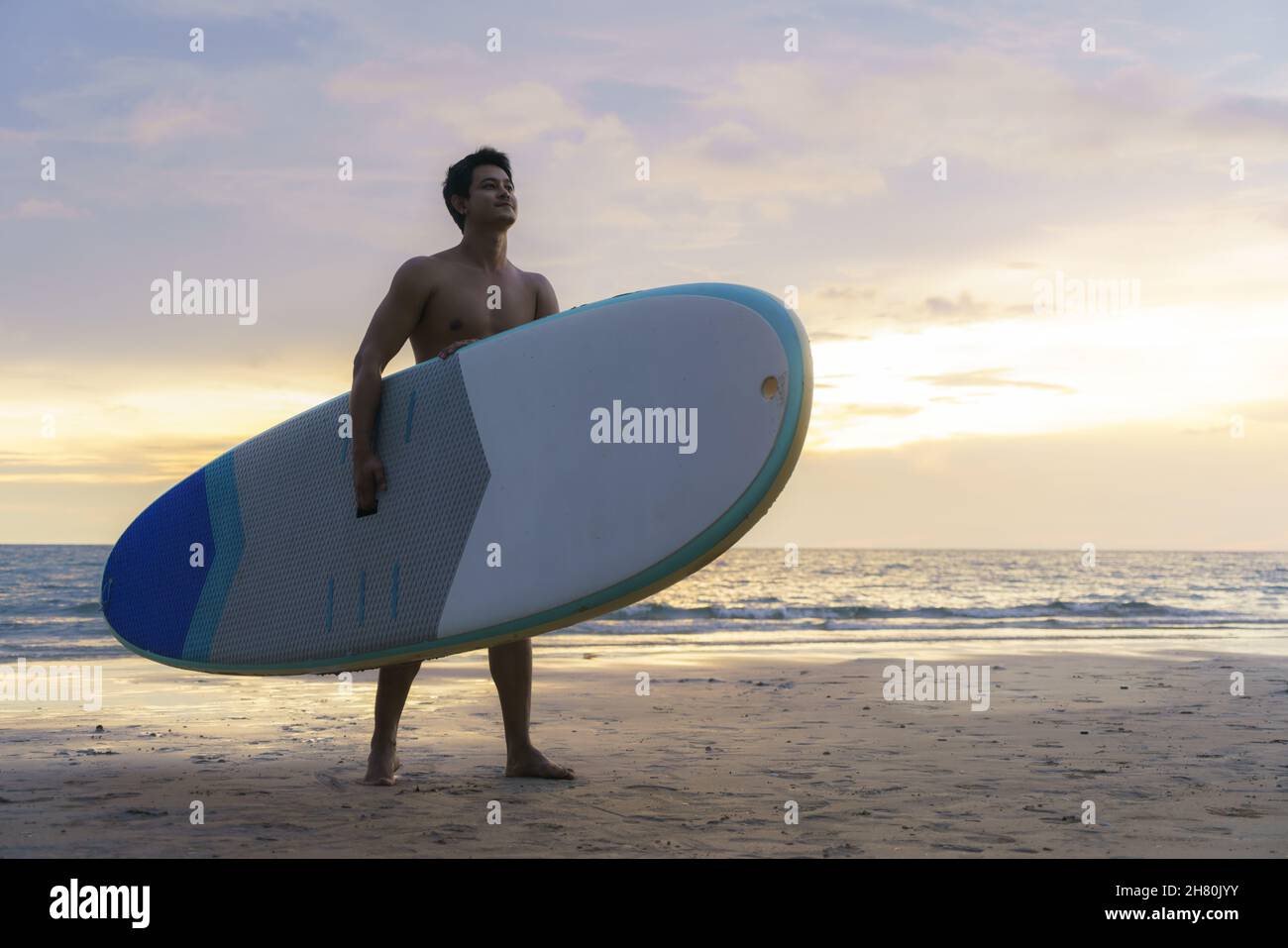 Asian man holding a sup board and paddle and walking on the beach. In the background, the ocean and the sunset. Back view. Summer surfing. Stock Photo