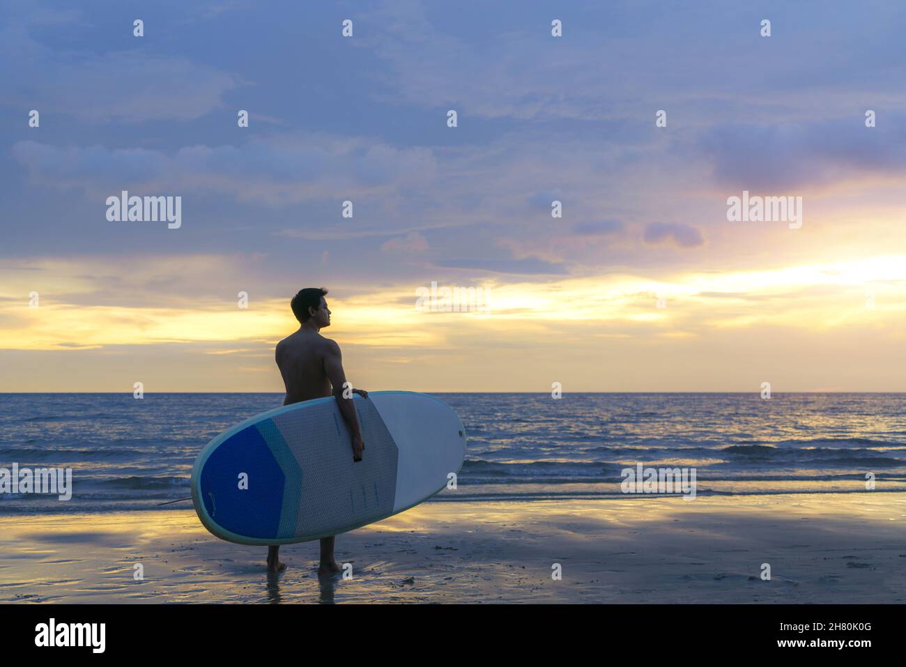 Asian man holding a sup board and paddle and walking on the beach. In the background, the ocean and the sunset. Back view. Summer surfing. Stock Photo