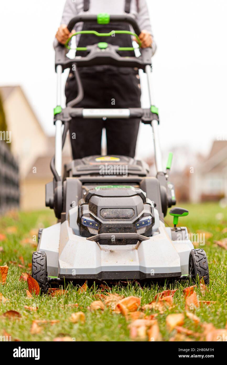 caucasian woman wearing boots and overalls is mowing the lawn in late fall. She is operating an electrical walk behind device. She goes over fallen le Stock Photo