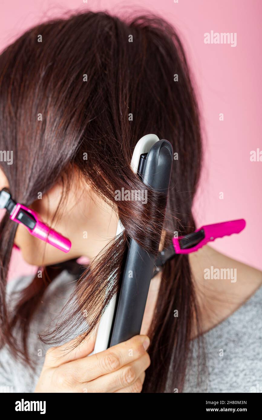 A beautiful young caucasian woman is using sectioning clips and heat hair iron wand for straightening or curling her hair. She is against pink backgro Stock Photo