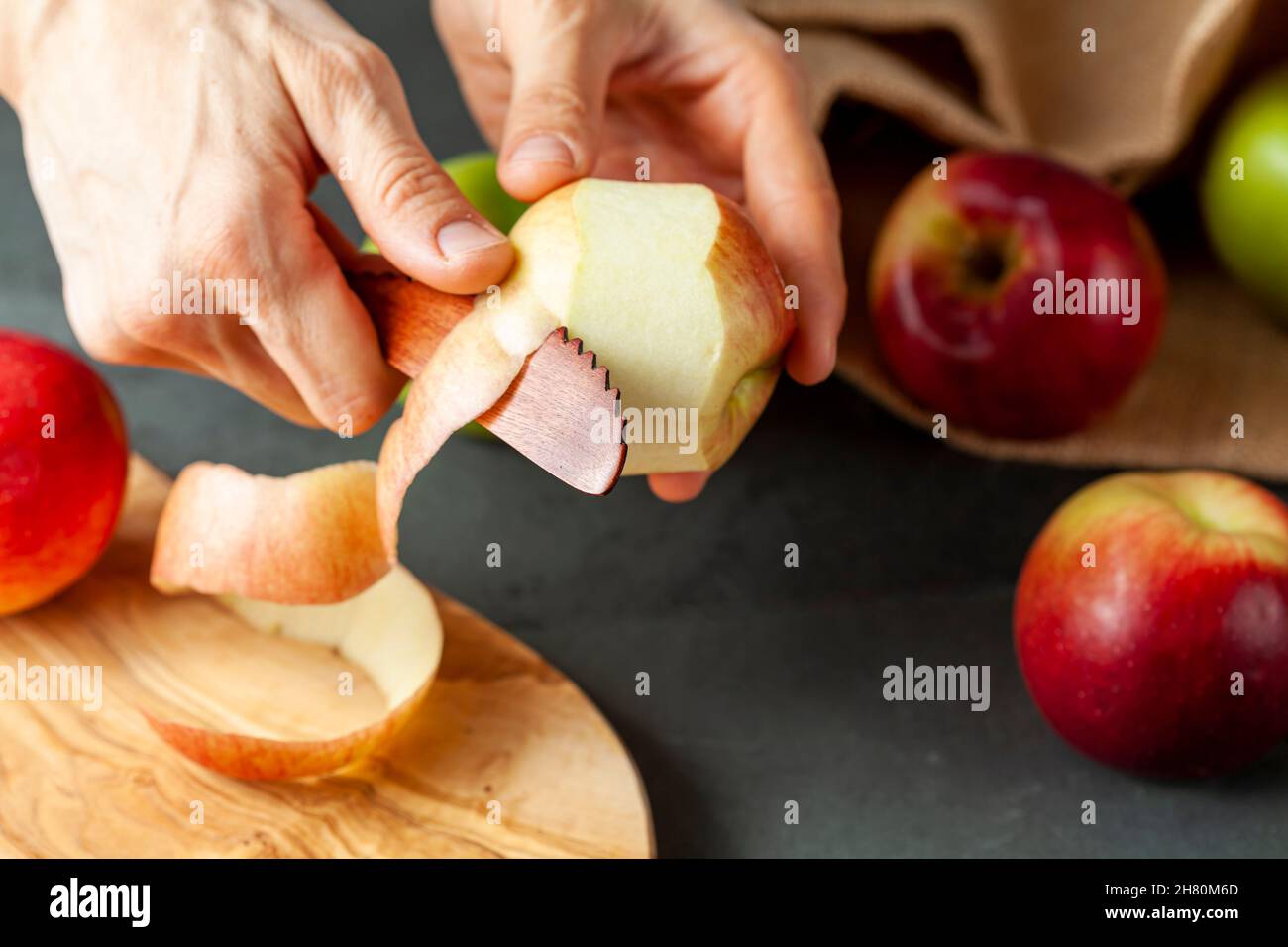 apples of different varieties are seen on dark stone and wooden background.  A woman is peeling the skin of an apple using a fruit knife. Healthy eati Stock Photo