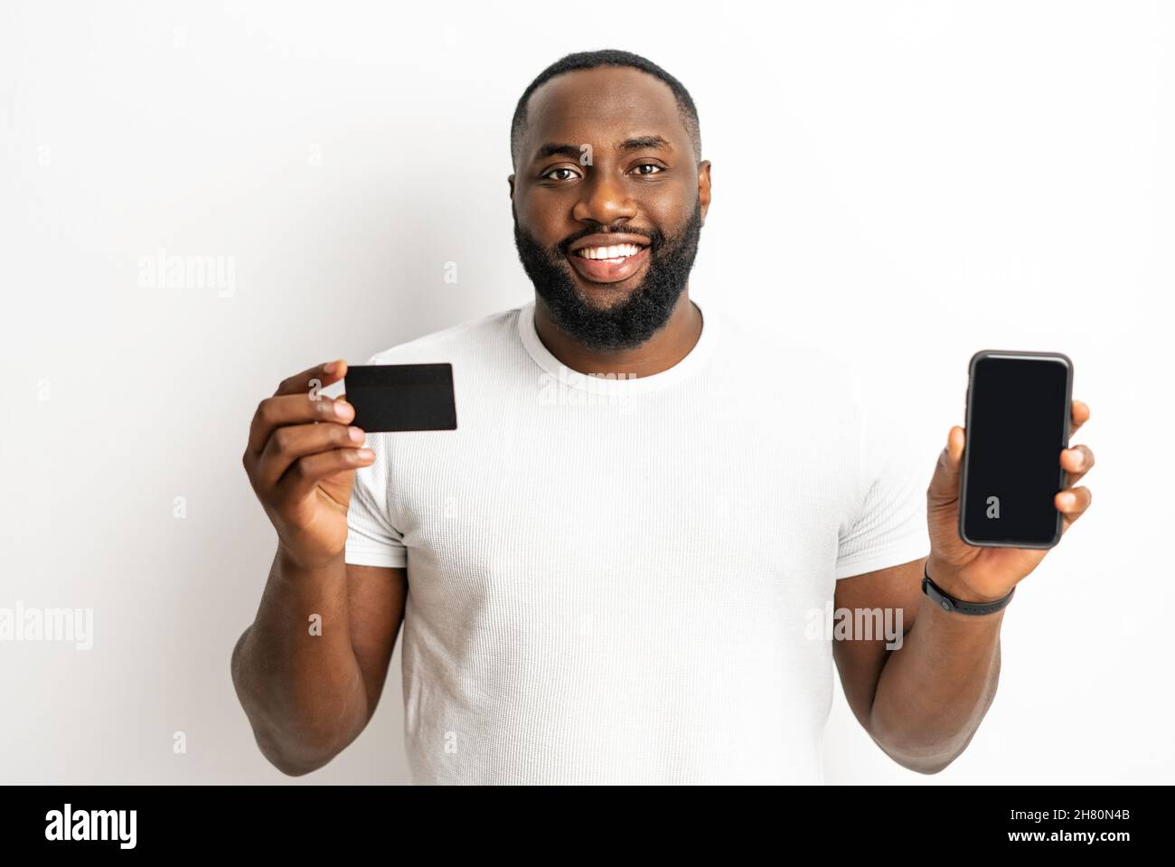 New mobile banking app. Smiling young African-American holding debit card and smartphone with empty screen isolated on white. Happy black man in casual t-shirt is advertising, copy space, mock-up Stock Photo